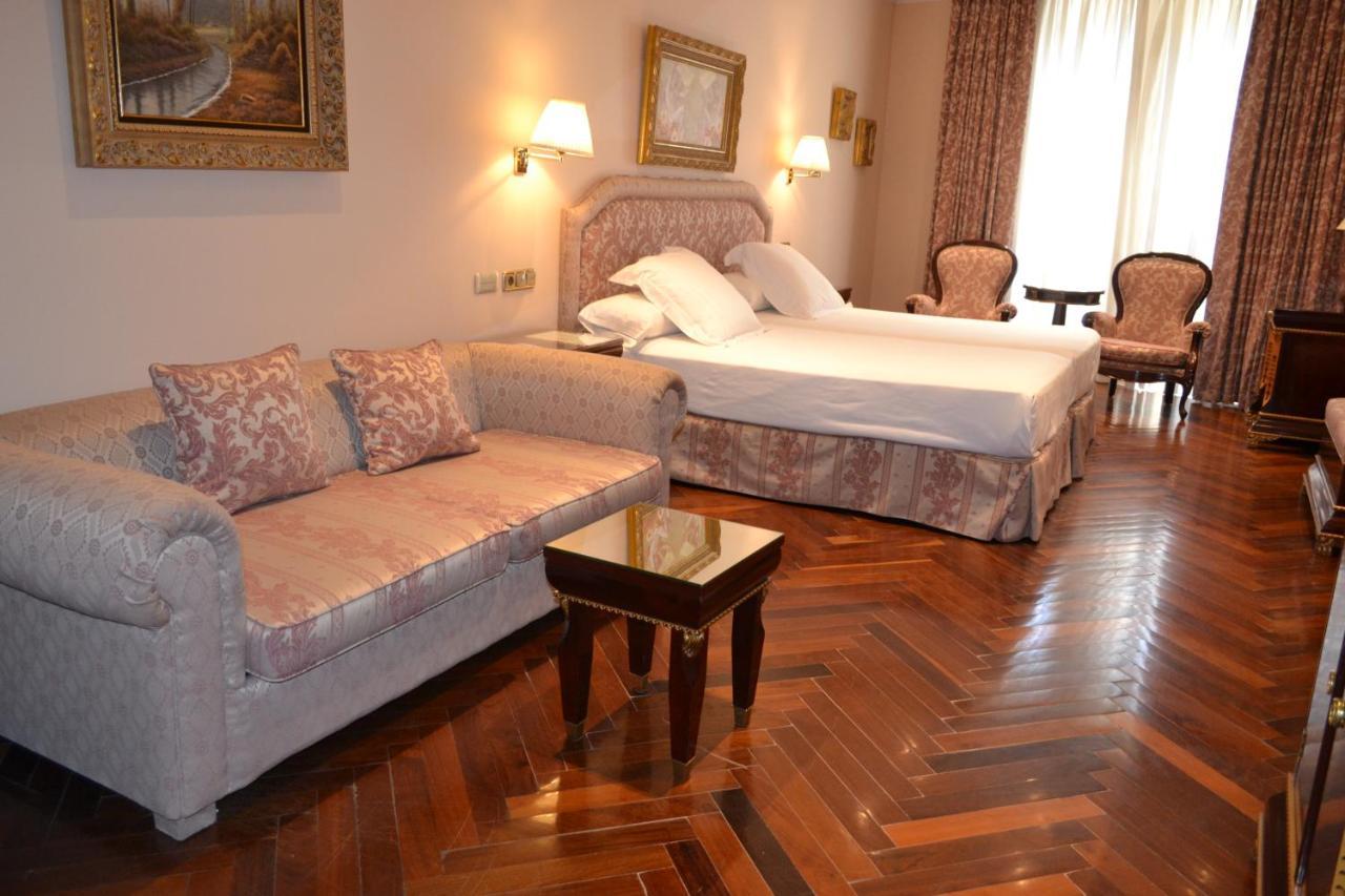 Hotel Alameda Palace Salamanca Kültér fotó The photo shows a hotel room featuring a queen-sized bed with white linens and a decorative headboard. There is a light-colored sofa with decorative cushions positioned to one side, accompanied by a small coffee table. On the opposite side, there are