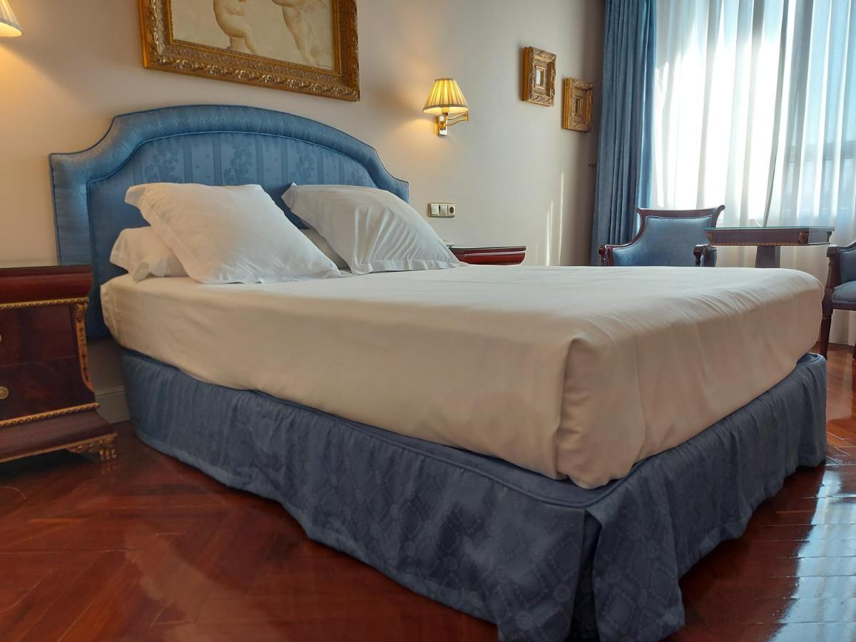 Hotel Alameda Palace Salamanca Kültér fotó The photo shows a well-furnished hotel room featuring a large bed with a light-colored mattress and decorative pillows. The bed has a soft blue headboard and a matching blue bed skirt. Beside the bed, there are bedside tables with lamps. In the backg