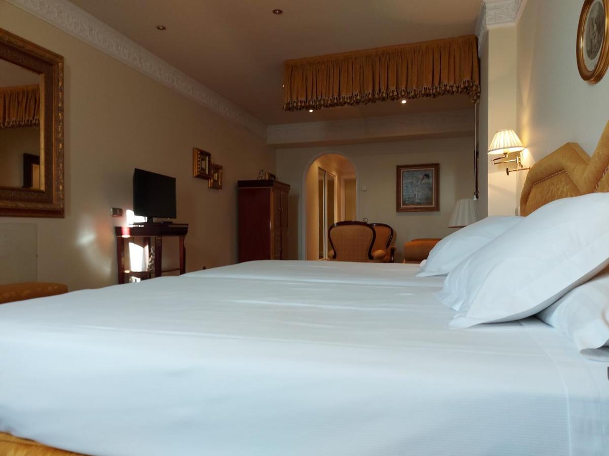 Hotel Alameda Palace Salamanca Kültér fotó The photo shows a hotel room featuring two beds with white linens. In the background, there is a mirrored wall that adds to the room's spacious appearance. To the left, a TV is mounted on the wall, and there’s a wooden cabinet underneath it. On the r
