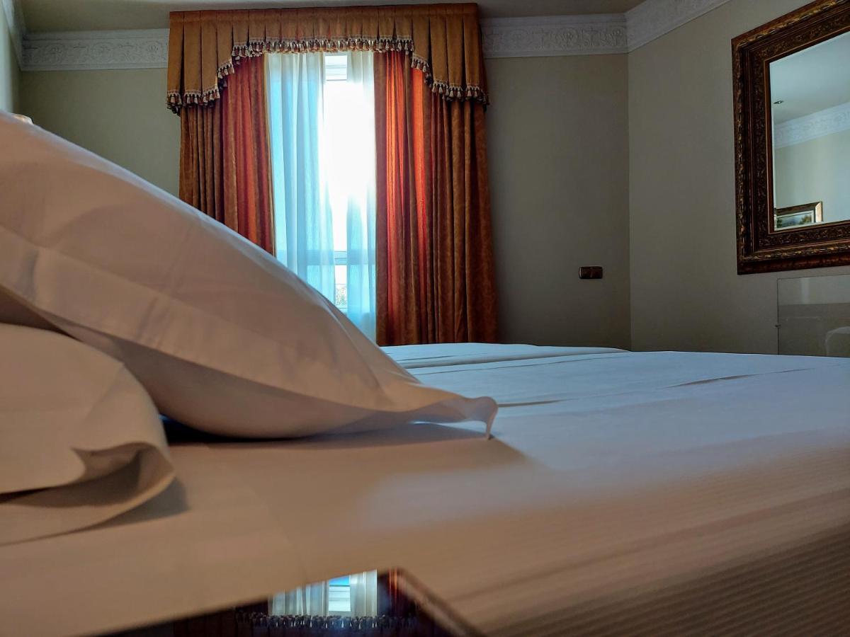 Hotel Alameda Palace Salamanca Kültér fotó The photo shows a close-up view of a bed. There are several white pillows placed on the bed, which has a neatly arranged white bedspread. In the background, there's a window with sheer curtains and darker drapes, allowing light to filter through. A m