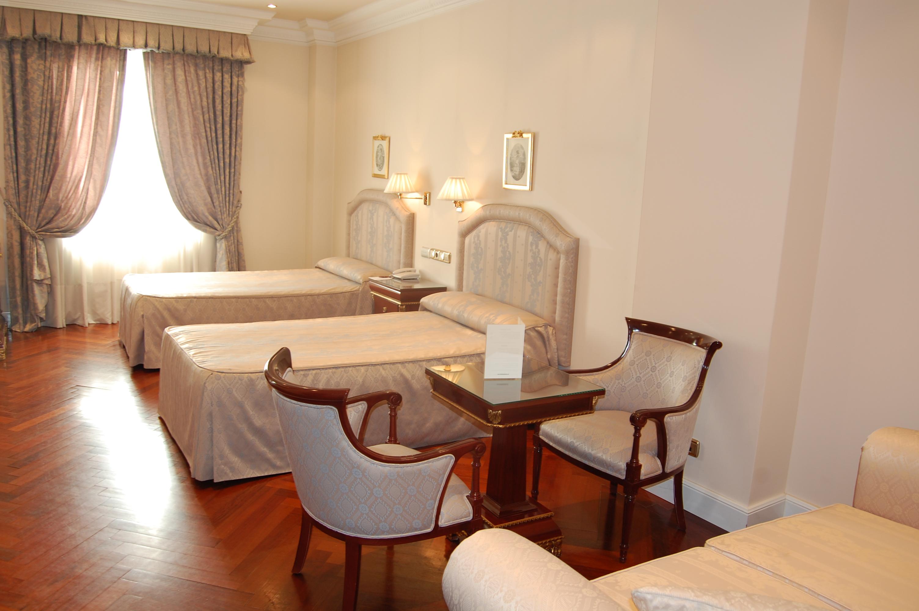 Hotel Alameda Palace Salamanca Kültér fotó The photo shows an interior of a hotel room featuring two single beds with light-colored bedspreads. There are curtains over a window allowing natural light to enter the room. A small table is positioned between two upholstered chairs, creating a coz