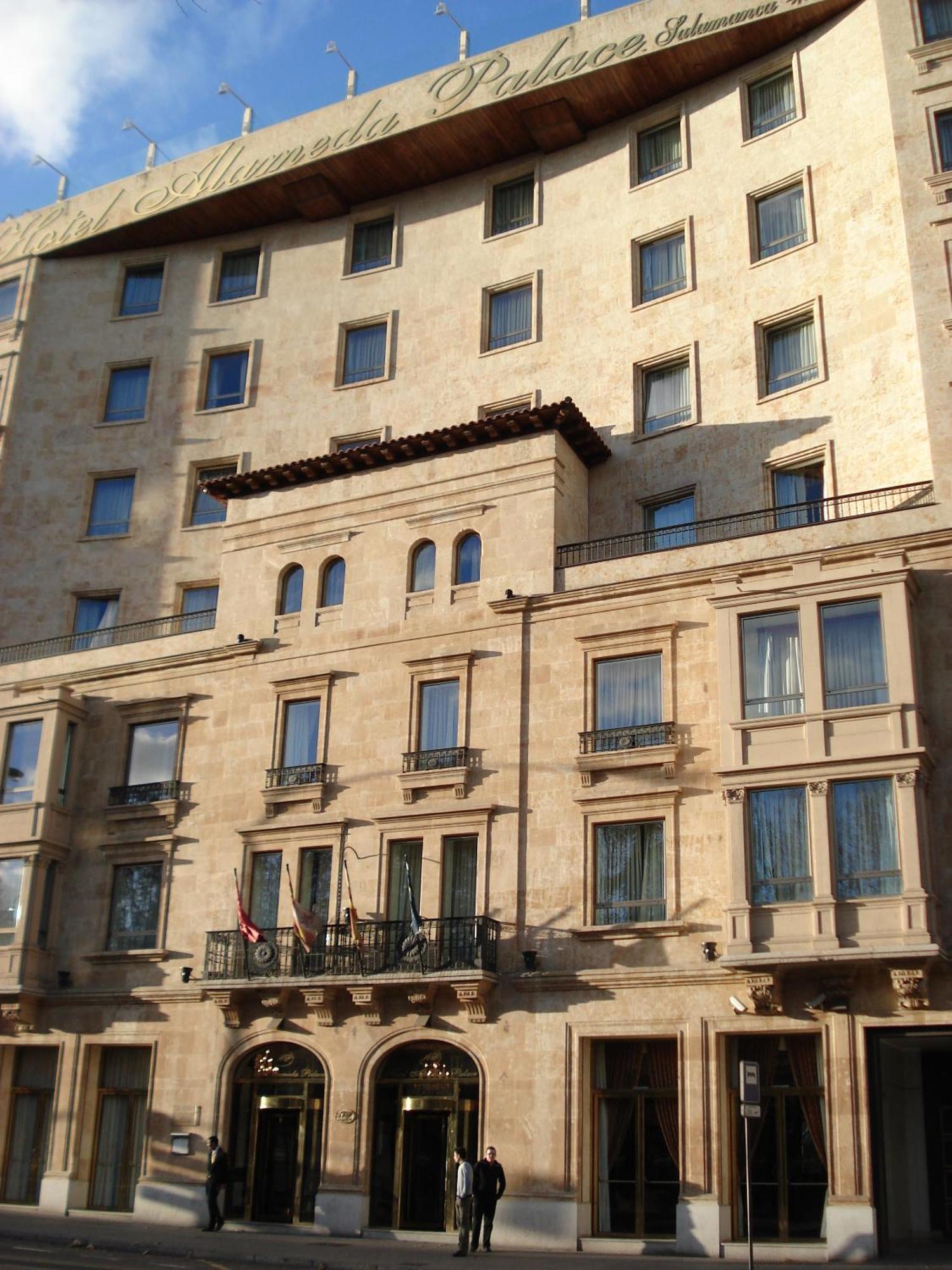 Hotel Alameda Palace Salamanca Kültér fotó The photo depicts a building with a stone facade that features a mix of architectural designs. The lower section includes several large windows framed by decorative elements, with balconies that have railings. There are also flags visible at the entr