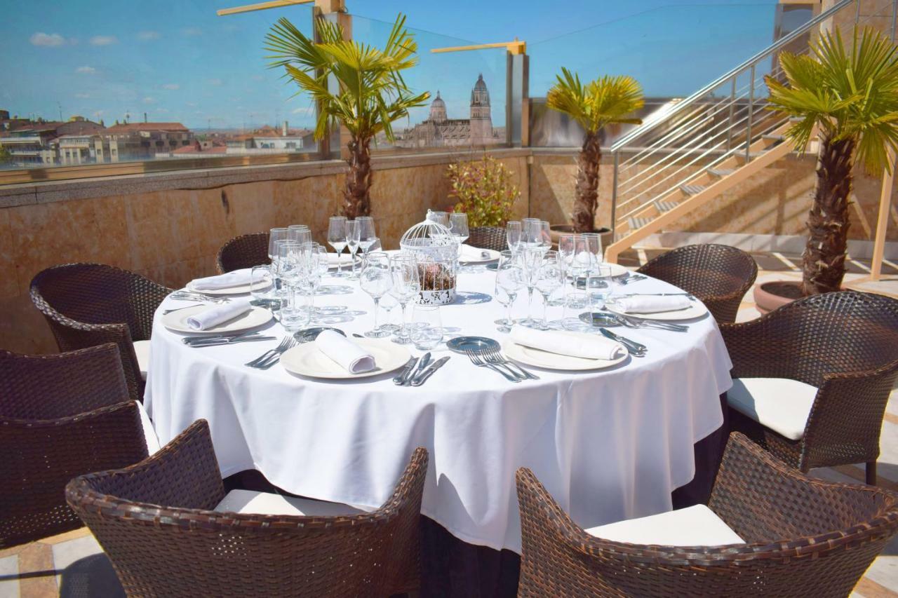 Hotel Alameda Palace Salamanca Kültér fotó The photo shows a round dining table set for a meal, covered with a white tablecloth. The table is surrounded by wicker chairs and features several glassware items, including wine glasses and water glasses, as well as cutlery and napkins neatly arran